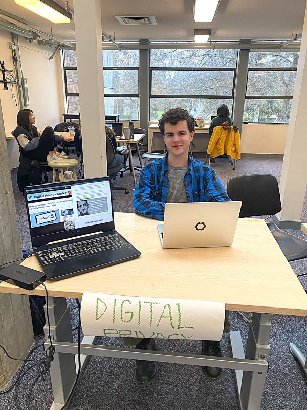 a student in a blue button down smiles at the camera, with their laptop open and visible beside them. There is a poster on the table facing the camera that says "Digital Privacy".