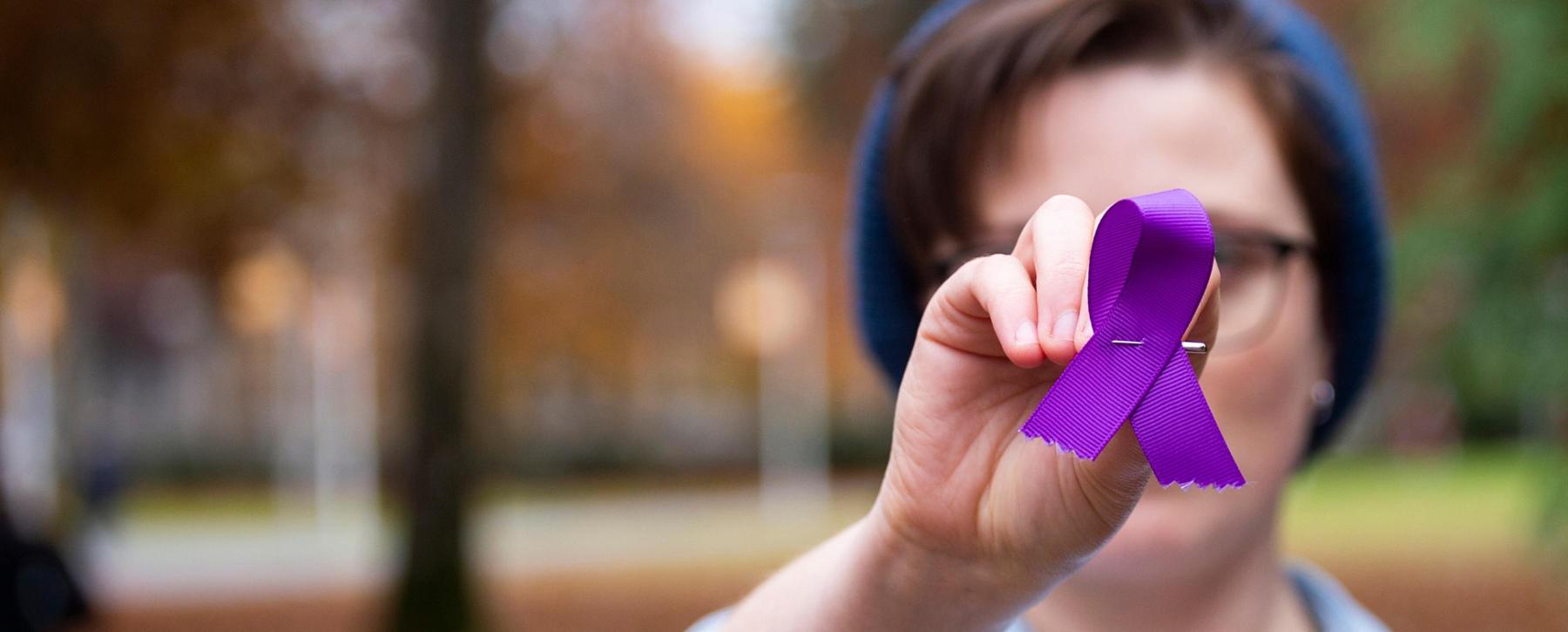 Hand holds purple ribbon in foreground. Background is blurred.