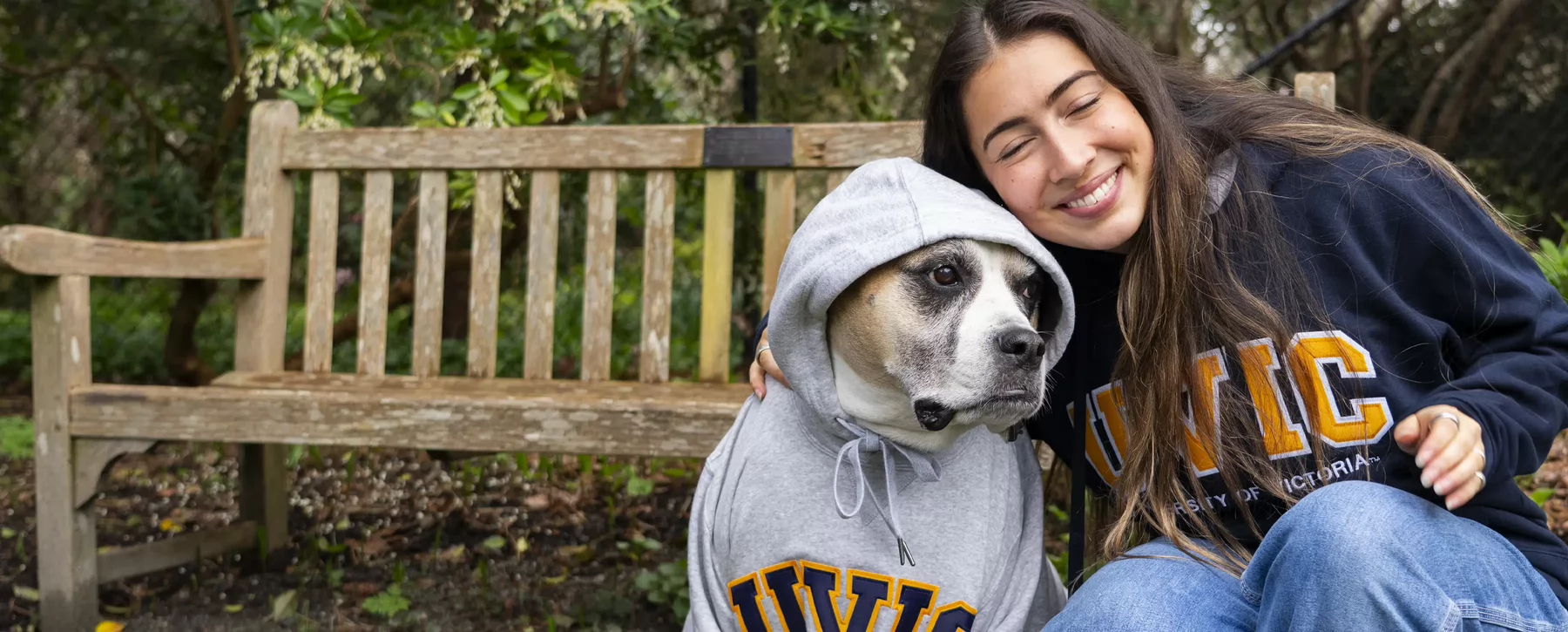 A student hugs a dog that is wearing a UVic hoodie.