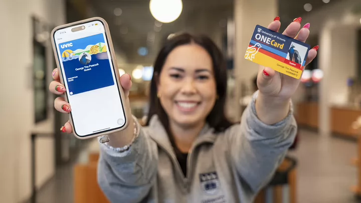 A smiling student holds up a mobile phone with a digital ONECard displayed and a physical ONECard. 