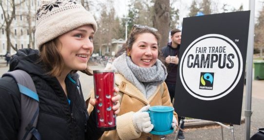 Two students stand beside UVic's Fairtrade campus designation sign while drinking coffee from reusable mugs.