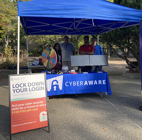 picture of cyberaware booth in front of the UVic quad. Four people are standing behind the booth and a prize wheel is on the booth table.