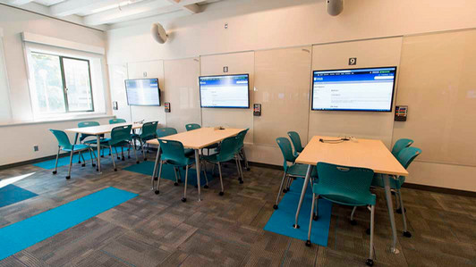 Three pods in MAC D115, each one with a large TV, a table, and 5 turqoise plastic chairs. There are glass surfaces on the walls between the pods and whiteboard pens in a holder. There's also a exterior window which has blinds and a handle to open.