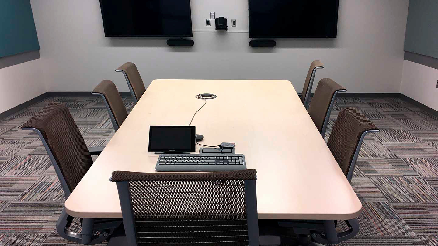 A small conference room with a long table and six chairs. There's a keyboard, mouse, touchscreen panel, and mic array on the table. The back wall has 2 large TVs and some other electronic equipment mounted to it.
