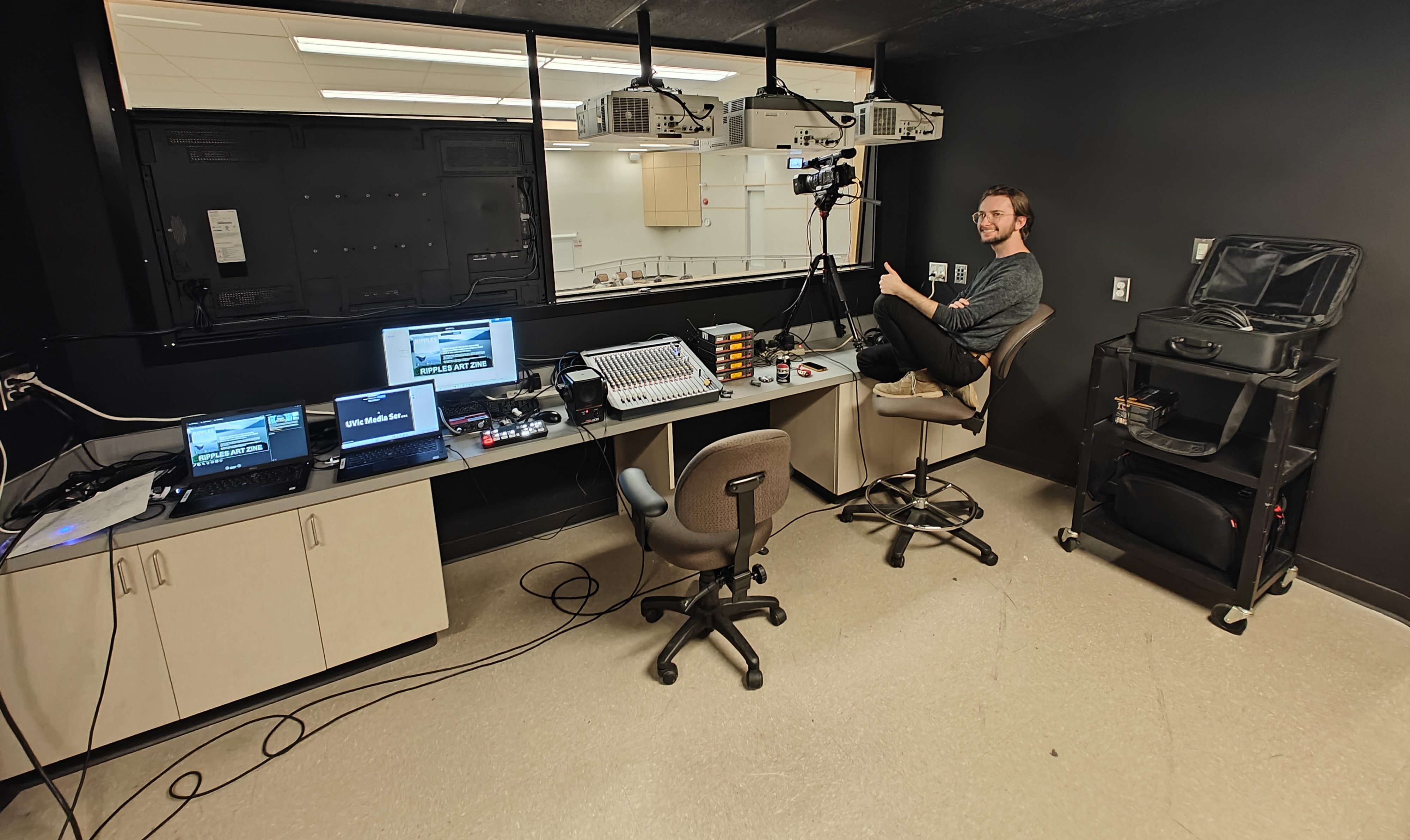 A cheerful AV technician making a thumbs up gesture while recording video from the projection booth of a lecture theatre.
