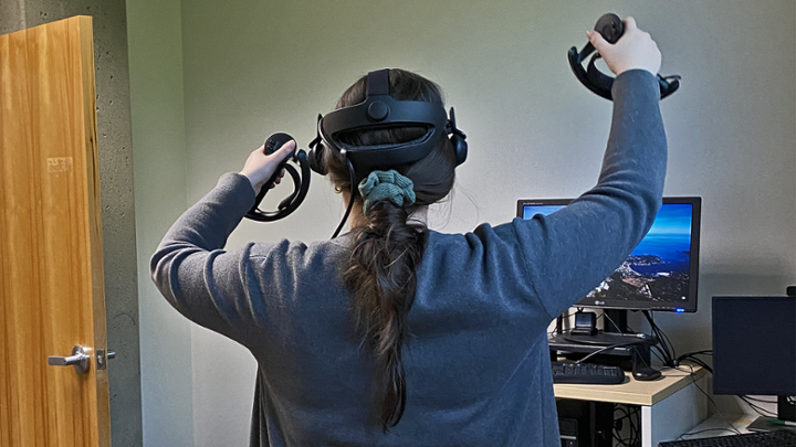 A student wearing a virtual reality headset, holding game controllers