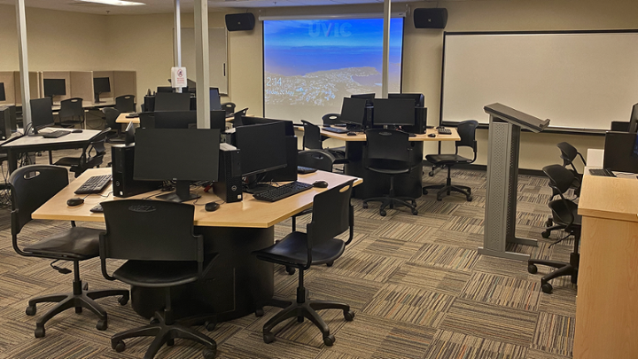 Classroom with computer tables in centre, some computers around edge of room facing wall.