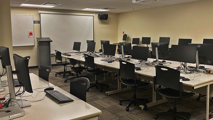 Classroom with computer tables in centre, some computers around edge of room facing wall.