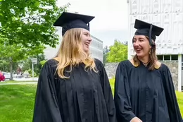 Two students wearing black graduation gowns and caps smile and talk while walking outdoors on a university campus. Trees, grass, and a modern campus building are visible in the background.