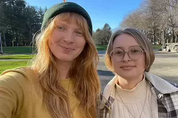 Two people smile for a selfie outdoors on a sunny day; one wears a green knit hat and yellow top, while the other wears round glasses, a cream sweater and a plaid jacket.