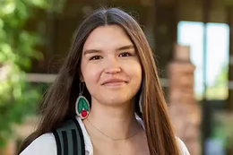 A young woman with long dark hair, wearing a backpack and a beaded earring, stands outdoors with trees and a building in the background.