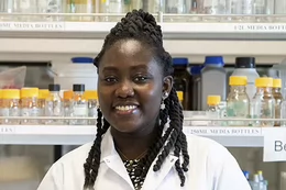 Smiling woman in lab coat stands in front of shelves of glass bottles