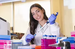 A woman in a white lab coat and blue gloves smiles while using a pipette in a laboratory. She is surrounded by lab equipment, including test tubes, racks, and containers in bright colours.