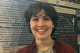 A smiling person with short dark hair wears a burgundy button-up shirt over a grey T-shirt, standing in front of an academic poster about queer literature.