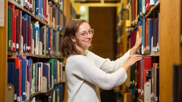 A young woman smiles at the camera as she reaches for a book in the library