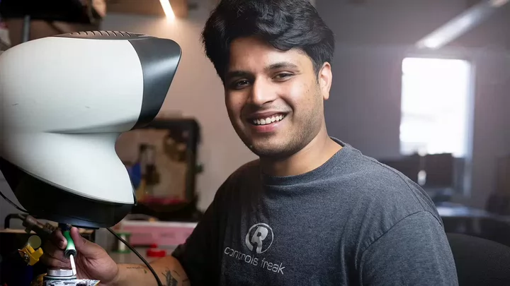 A smiling person sits at a workstation using a large piece of technical equipment, wearing a t-shirt that says “controls freak.”