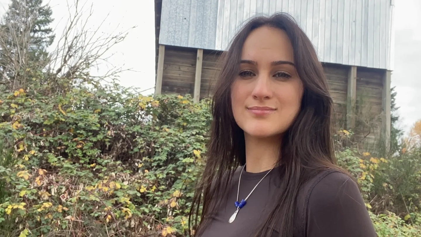 Young woman standing outdoors in front of greenery and a wooden building, wearing a dark top and a necklace.