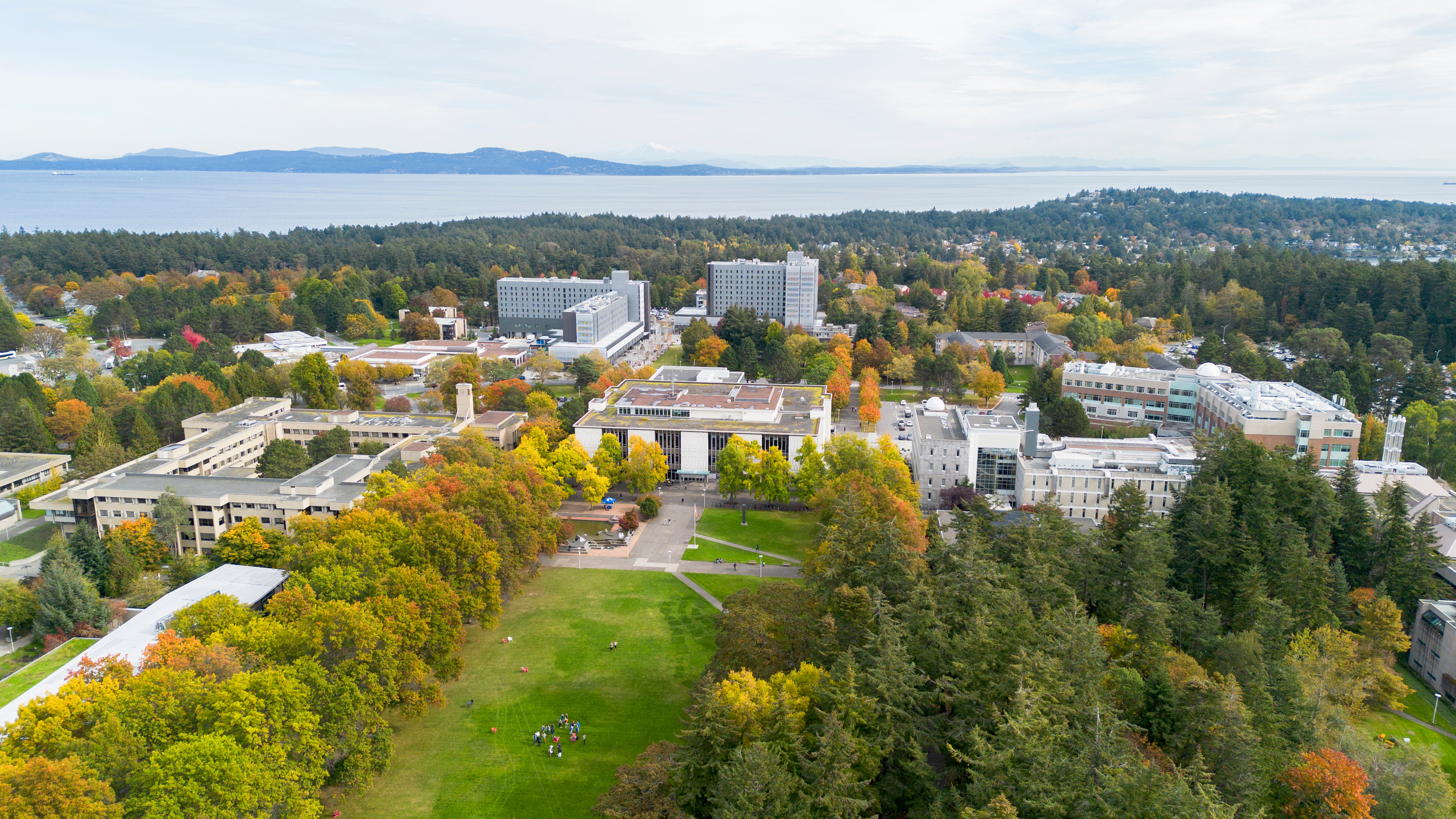 An aerial photo of a university campus surrounded by trees, with ocean and mountains in the distance.