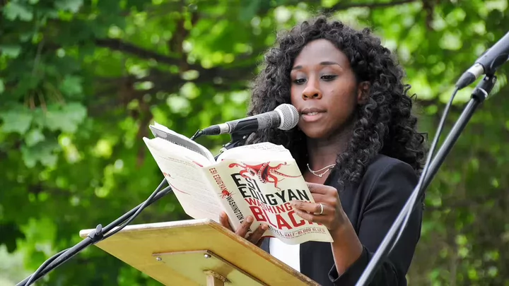 A woman reads from an open book at a podium with a microphone during an outdoor event, with trees in the background.