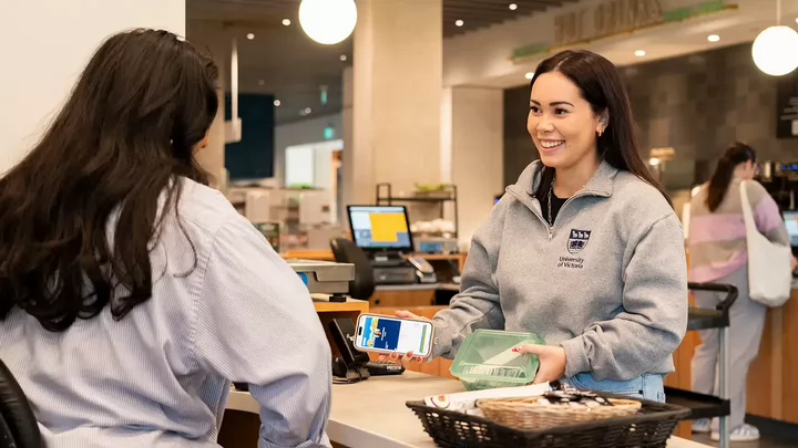  A person with long brown hair, wearing a gray University of Victoria sweatshirt, smiles as she pays for a meal with her phone. She is holding a green reusable to-go container and talking with a cashier.