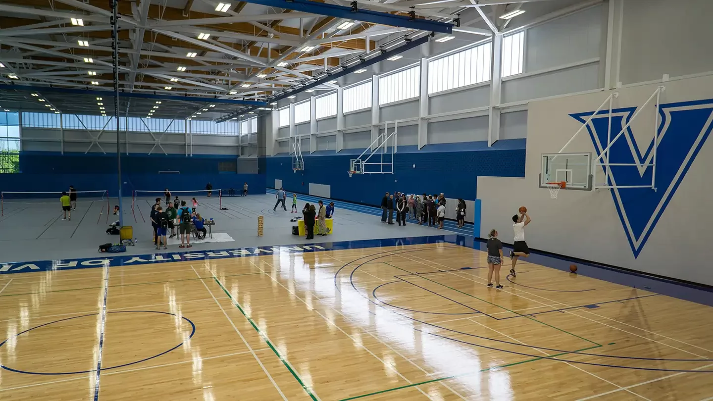 Wide interior view of a large indoor gymnasium with multiple marked courts, showing people playing basketball on the right, others gathered around tables and activities in the centre, and additional sports equipment set up across the floor under a high ceiling with skylights.