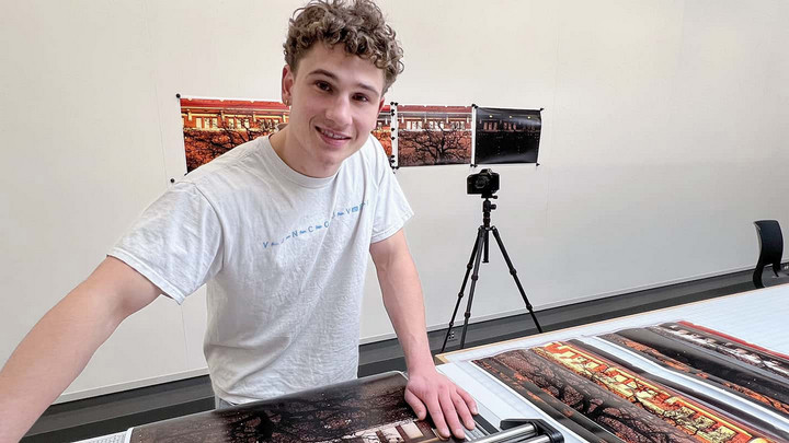 Student working in a photo lab cutting large prints to size. Some photo prints pinned to the wall behind him.
