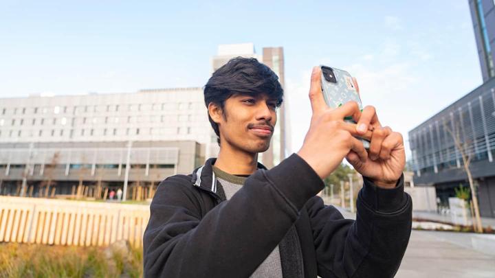 Stave standing outside residence buildings on campus taking a photo with his phone