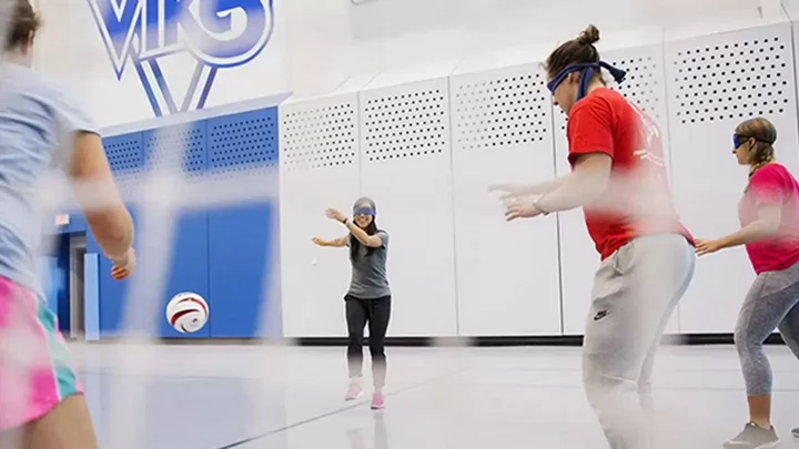 Students wearing blindfolds participate in an indoor goalball-style activity, moving across a gym floor toward a ball during an adaptive sport session.