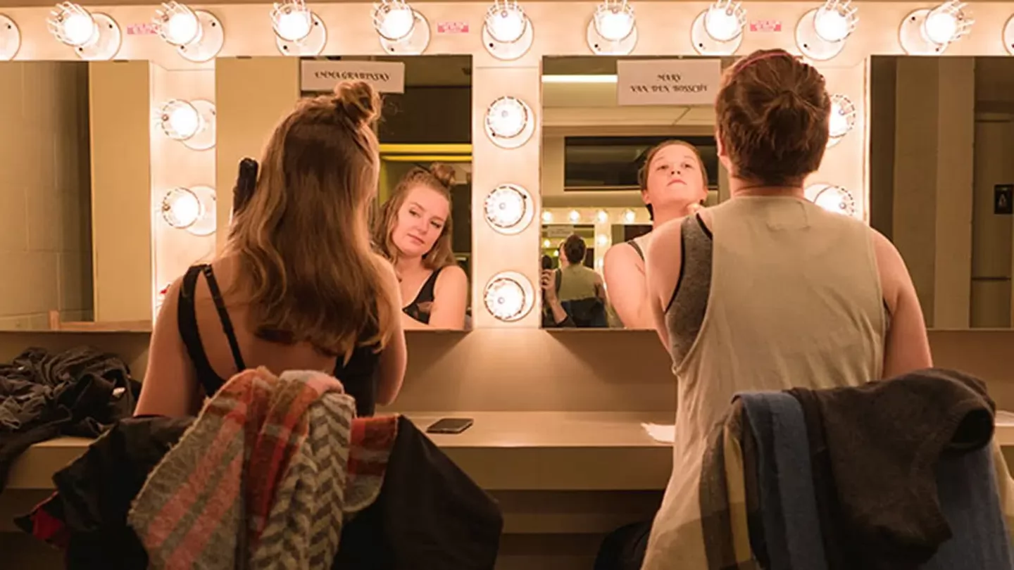 Two theatre students sit at a backstage dressing room counter, facing illuminated mirrors as they prepare for a performance.