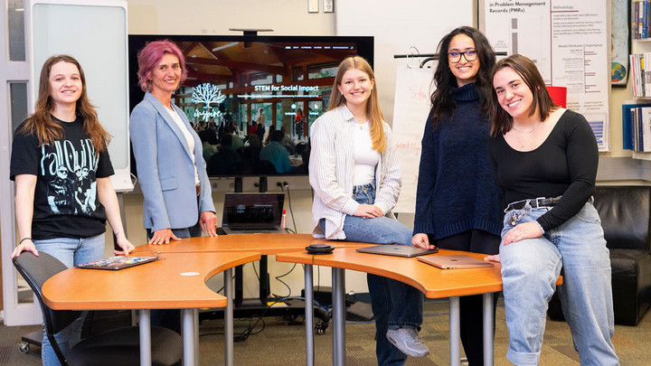 Valeriya Savchenko, Daniela Damian, Remee Brown, Kezia Devathasan, Shyla Burns sitting and standing around a table with a screen in the background showing the INSPIRE logo and the words, 'STEM for Social Impact"