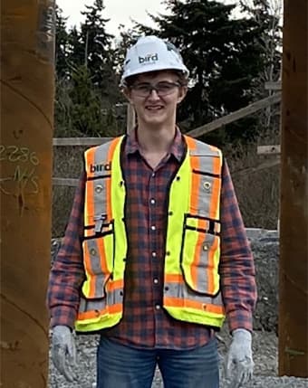Ethan Barillo wearing a hardhat, safety vest and glasses and work gloves stands in a work site with large metal pillars on each side