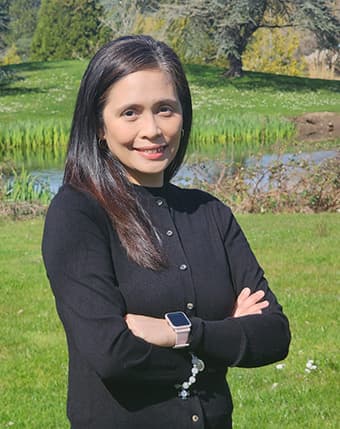 Nina Kanapi wearing a black sweater fully buttoned to the top with her arms crossed standing outside on a lawn with a creek in the background