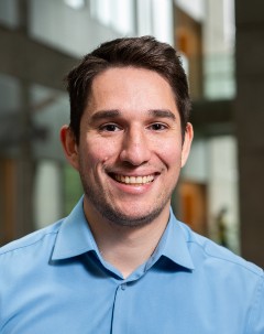 smiling man with dark hair wearing a blue, button-down dress shirt.