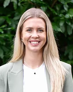A smiling woman with long blonde hair stands indoors wearing a light green blazer over a white top.
