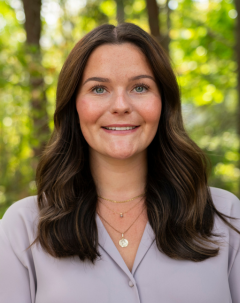 smiling woman with long brown hair, white blouse with green foliage behind