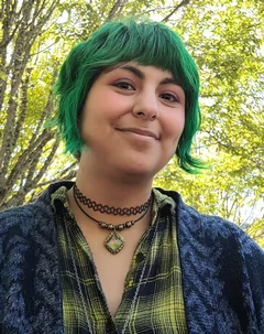 Person with short bright green hair smiling at the camera outdoors beneath leafy trees, wearing a green-and-black plaid shirt, dark blue cardigan and layered necklaces.