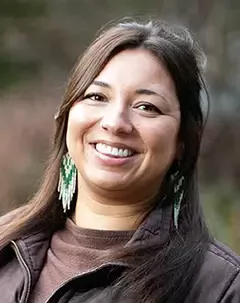 Smiling person with long dark hair wearing a brown jacket and green beaded earrings, photographed outdoors against a blurred natural background.