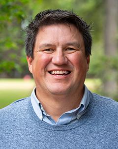 smiling man with dark hair and light blue sweater in front of a leafy green background